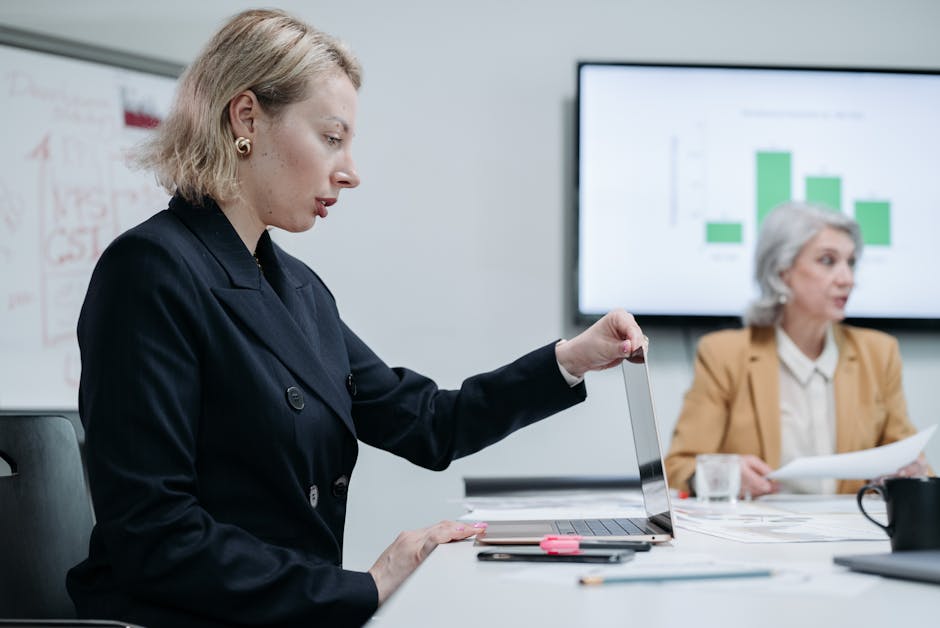 Two businesswomen discussing data in a modern office setting with laptop and charts.